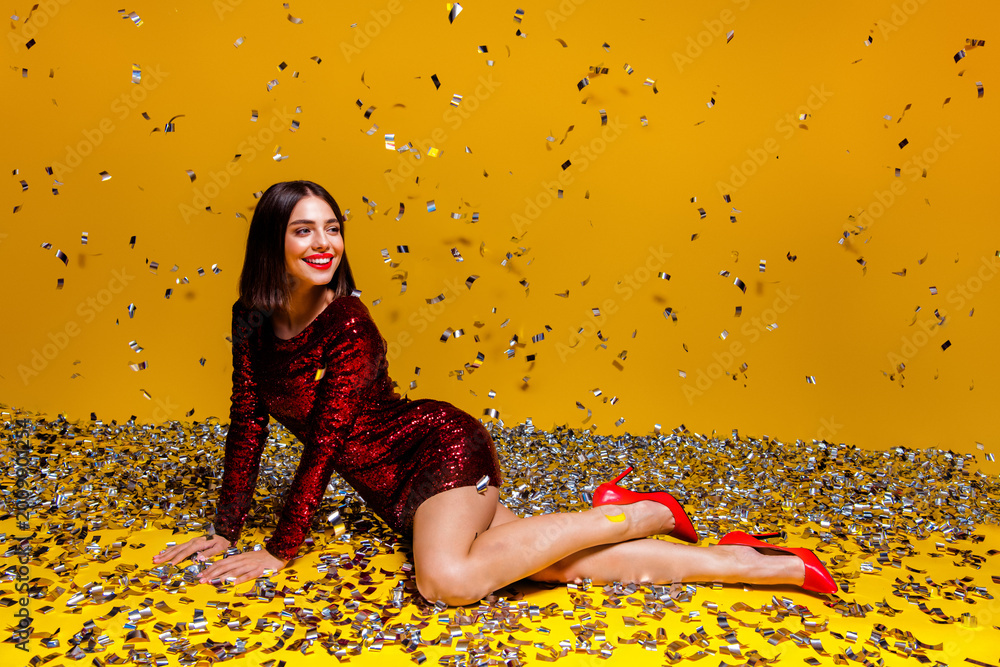 Naklejka premium Smiling woman in red sequin dress and high heels posing with falling confetti on yellow background for festive party celebration and glamorous studio portrait