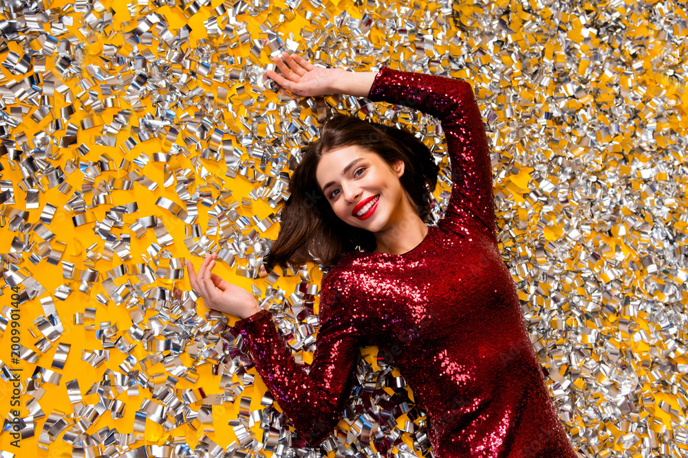 Fototapeta premium Smiling woman in a red sequin dress lying in silver confetti for a festive party celebration and holiday glamour photoshoot