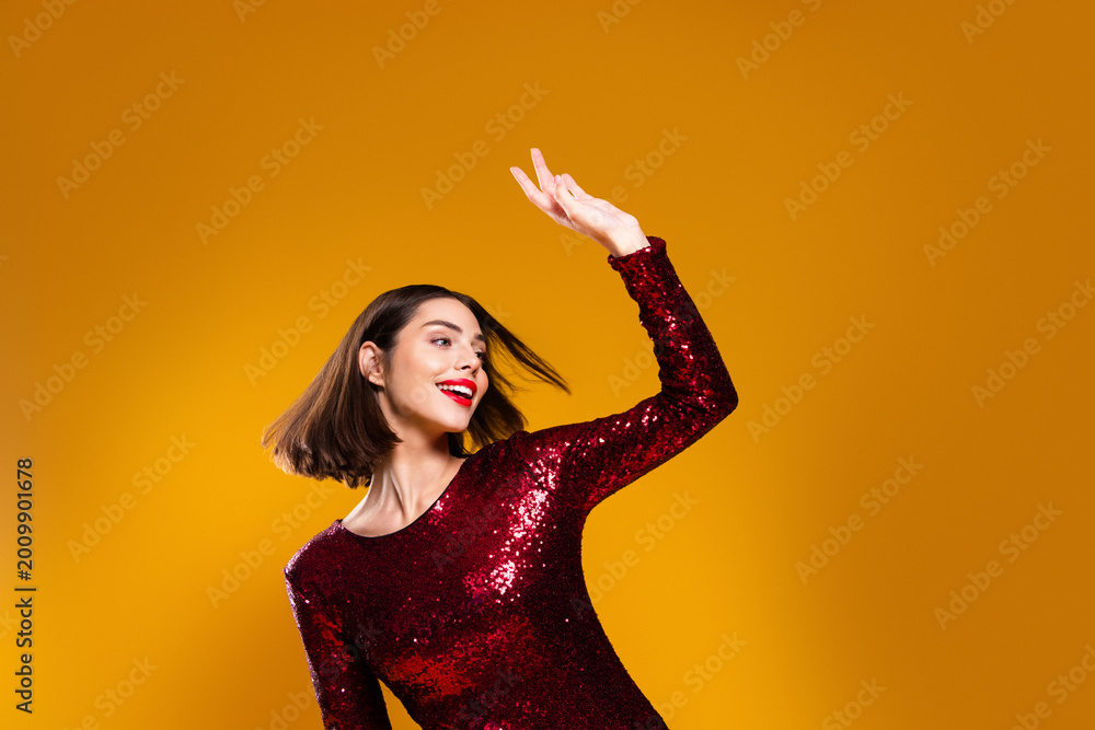Obraz premium Confident woman in red sequin dress dancing and smiling against a yellow background, glamorous party portrait conveying joy, energy and festive style