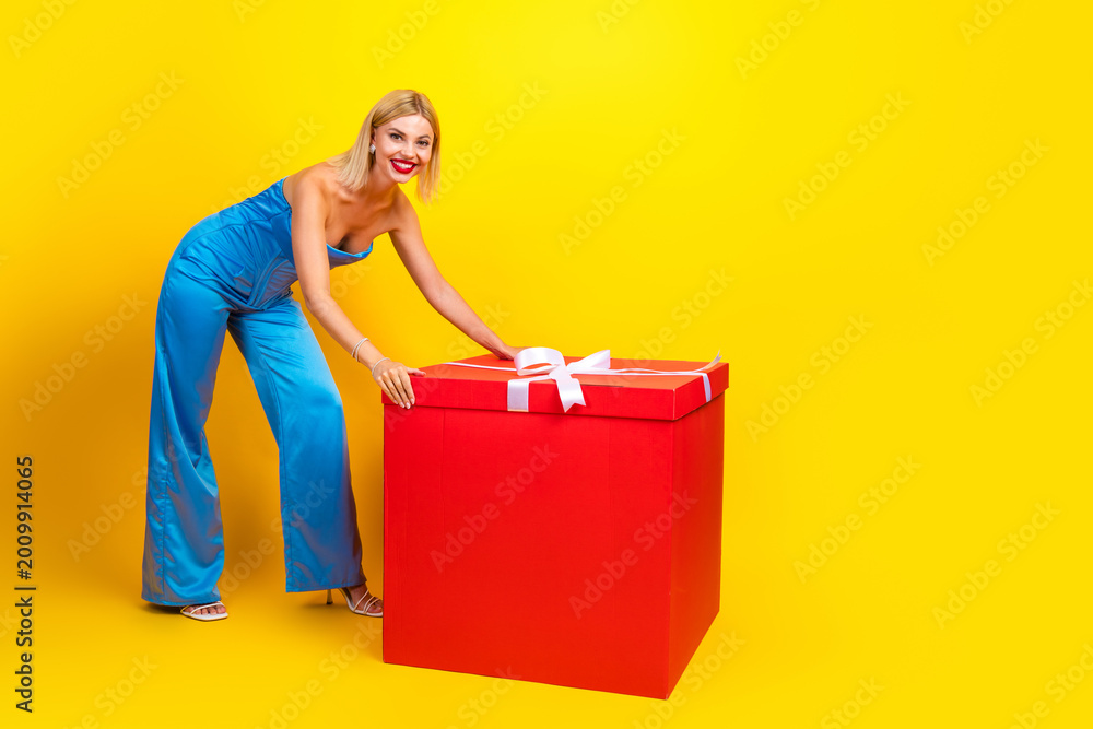 Obraz premium Young chic woman in blue jumpsuit greets a large red gift box against a bright yellow background
