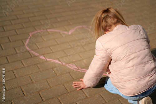 Young girl drawing pink chalk heart on outdoor pavement.