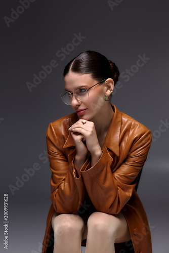 Canvas Print Stylish young woman in leather coat and glasses posing thoughtfully in a studio