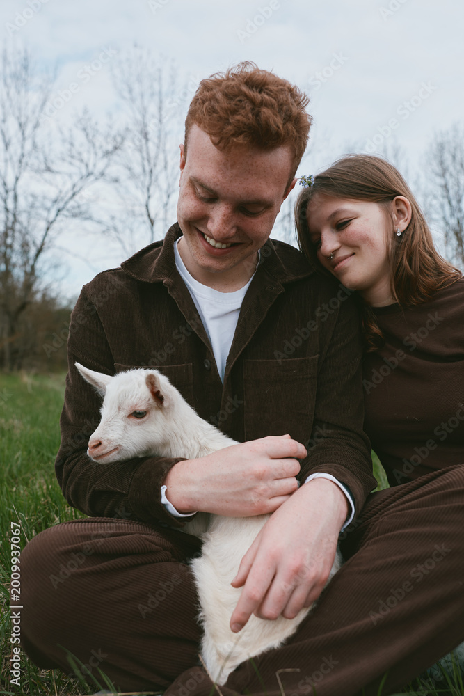 Fototapeta premium A couple shares a gentle moment with a cheerful goat kid in a grassy meadow, embracing outdoors, showing rural lifestyle and affectionate bond, serene skies, casual clothing, warm interaction,