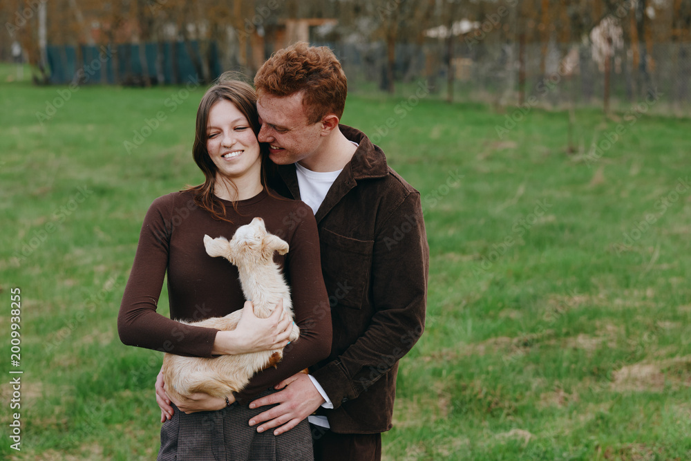 Naklejka premium A joyful couple shares a moment with a small goat kid in an open grassy park, embracing and smiling. A warm lifestyle scene that captures affection, companionship, and simple countryside moments.