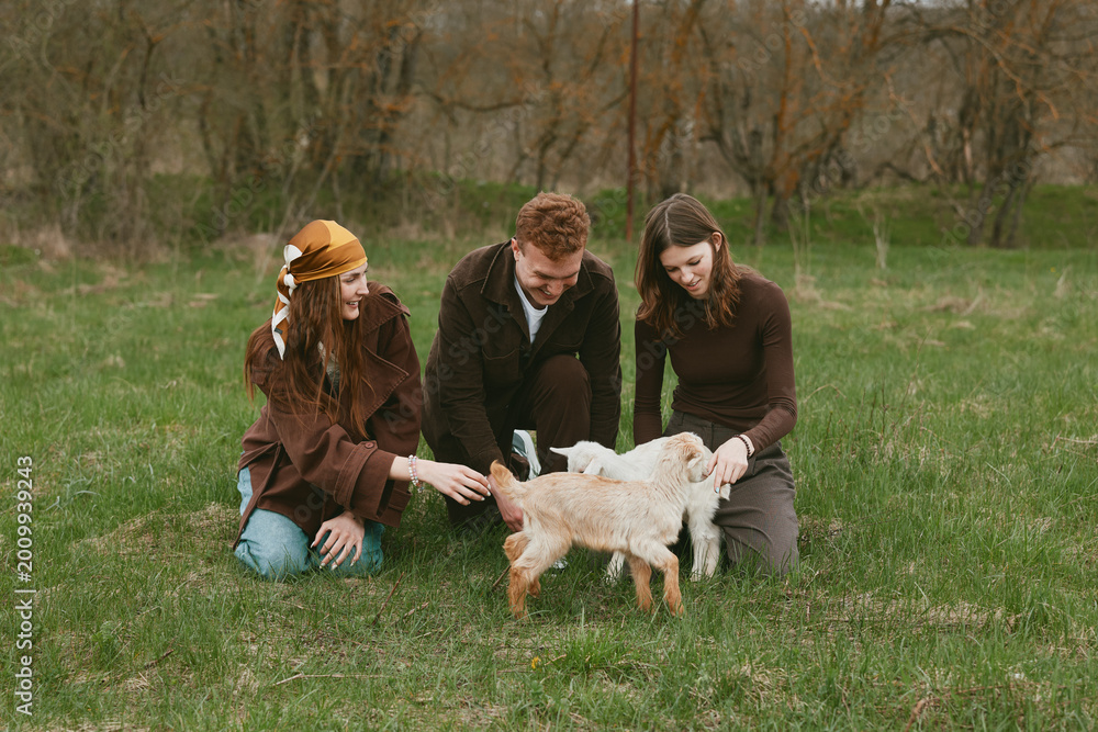 Fototapeta premium Goat kid animal family outdoor field nature interaction three people gently guide a playful goat kid in a grassy field, capturing a warm, candid moment of care and connection
