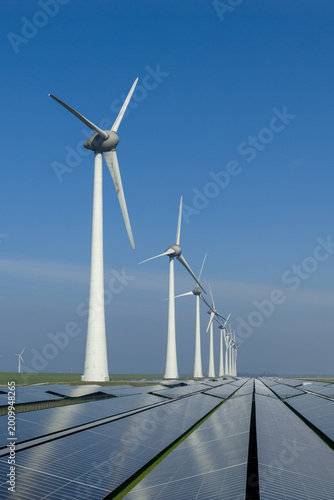 Aerial view of a row of tall wind turbines integrated with a large-scale solar panel array under a bright blue sky in the Netherlands.