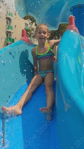 Happy child water slide under waterfall in childrens pool and laughs. Hotel water park. Summer vacation holiday travel. Vertical shot