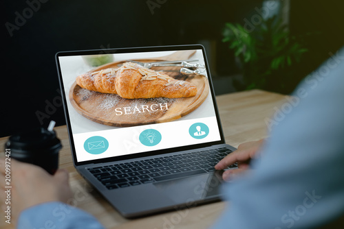 SEARCH Recruitment Selection Hiring job Person browsing a bakery website displaying croissants on a laptop while holding a cup of coffee at a wooden table.