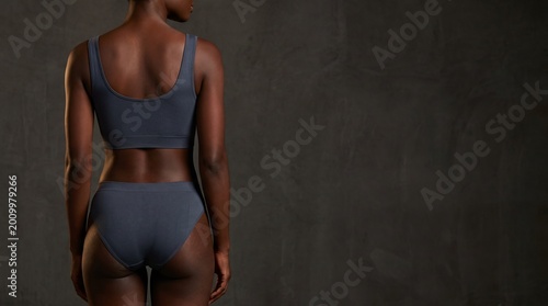 Back rear view close up cropped young black woman lady with athletic fit body dark skin she wear grey top bra underwear panties stand on dark textured wall background. Fitness wellness beauty.