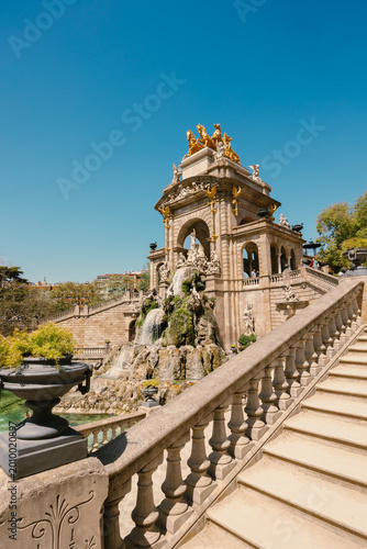 Low angle view of a grand stone fountain monument with cascading water, sculptures, a stone staircase, and a golden chariot statue.