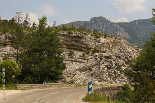 A picturesque winding road in the Alps against a backdrop of sheer cliffs and mountains. A mountain road in France: rock walls, pine trees, and clear skies. A trip through rocky terrain under the sun
