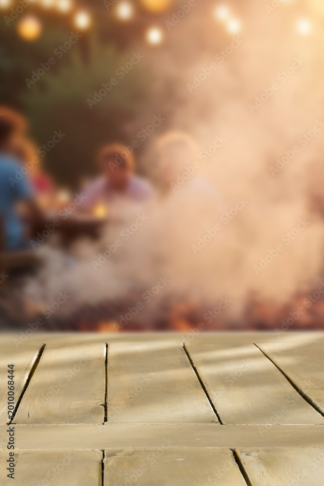Naklejka premium Empty wooden table with clean copy space for product placement in the foreground. Blurred background with garden camping scene with barbecue grill and people enjoying outdoor time, warm, relaxed.