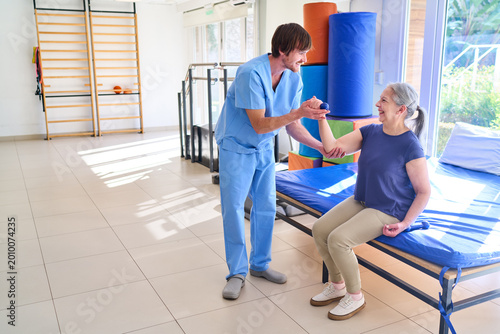 Senior Woman Lifting Dumbbell With Physiotherapist Assistance During Rehabilitation Session Indoors At Rehab Center For Recovery And Strength