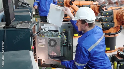 Industrial worker in blue uniform and hard hat inspecting robotic arms in a factory