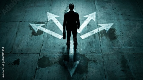 Man in suit standing at crossroads with multiple arrows on dark concrete floor representing career path choice opportunity Business decision and strategy concept