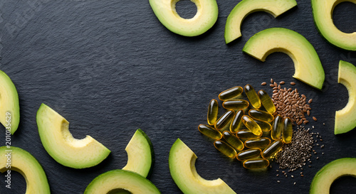 Arranged slices of fresh avocado alongside dietary supplements and seeds on a dark textured background
