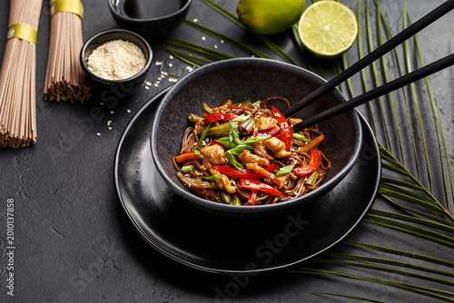Stir-fry soba asian noodles with chicken, vegetables and chopped green onion in ceramic bowl on black stone background with palm leaf, chopsticks, lime, soy sauce, sesame seeds and dry noodles