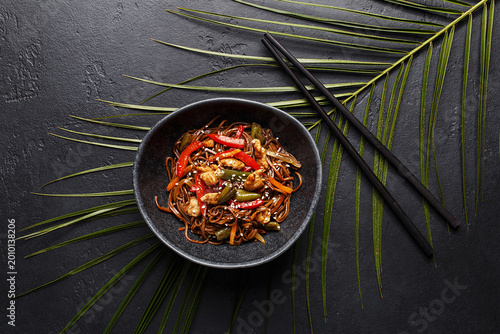 Stir-fry soba asian noodles with chicken, vegetables and sesame seeds in black ceramic bowl on black stone background with tropical palm leaf and chopsticks. Overhead view