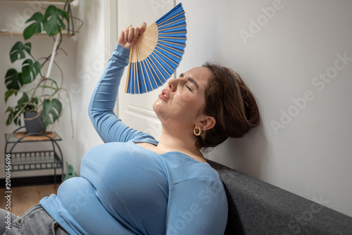 фотографія Exhausted overweight young woman on sofa suffering from overheat indoor using hand fan to cool down, breathing heavily