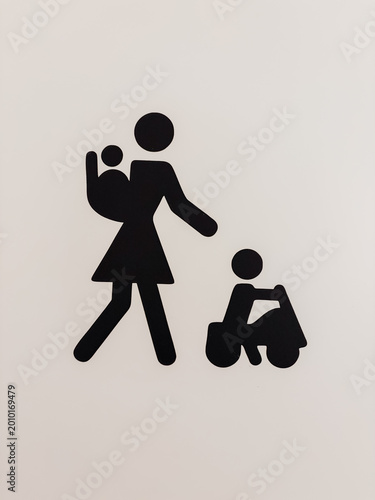Minimal black restroom sign showing mother with baby and child in toy car. Simple public facility symbol on clean white background.