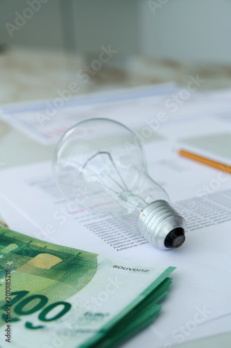 A light bulb lies on top of printed documents, with euros stacked close by on a desk in an office space.