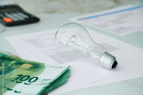Paperwork, a light bulb, cash, and a calculator are on a desk during a work session in an office.