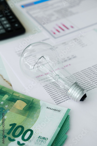 A light bulb lies on financial papers while a euro banknote is nearby, showing a workspace setting with a calculator.