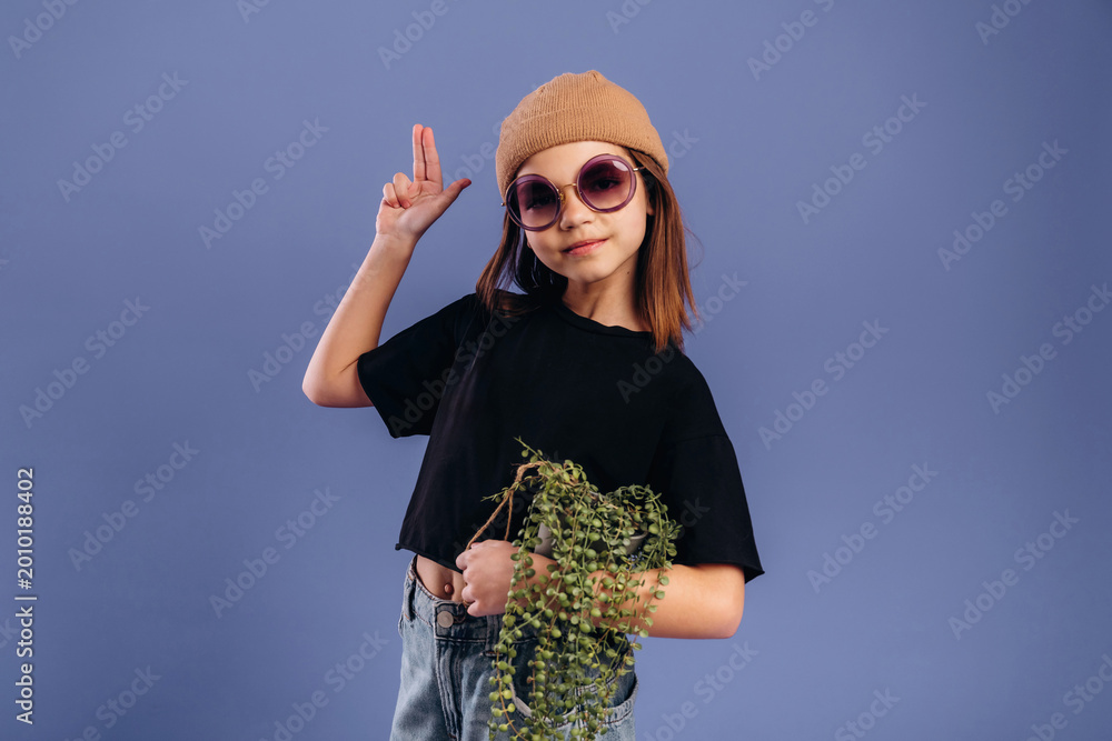 Naklejka premium Young girl with brown hair wearing oversized sunglasses and a beige beanie poses playfully while holding a potted plant against a solid purple background