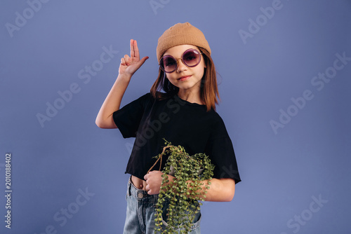 Young girl with brown hair wearing oversized sunglasses and a beige beanie poses playfully while holding a potted plant against a solid purple background