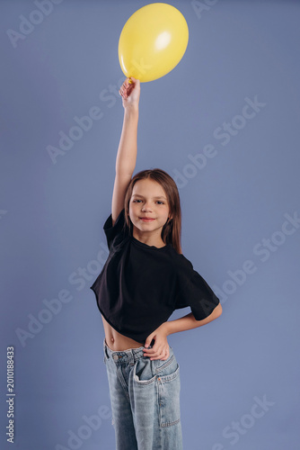 Young girl with long brown hair wearing a black crop top holds a yellow balloon high above her head against a solid blue background