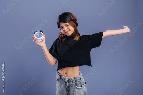 Young girl wearing black crop top and denim jeans holds alarm clock while stretching her arm against a solid blue background in a playful pose