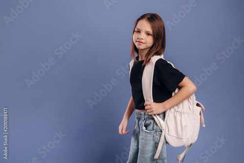 Young girl with brown hair wearing a black t-shirt and blue jeans carries a light backpack against a solid blue background, posing confidently for the camera