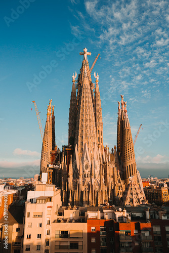 Rooftop view of the Sagrada Familia basilica towers at sunset with the central Jesus Christ tower and cross under a blue sky.