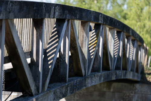 Close-up view of a wooden bridge railing with geometric support beams in a green outdoor park. Rustic footbridge detail with natural texture, shadows and shallow depth of field.