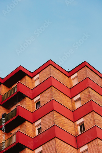Red and beige brick residential apartment building showing a modern zigzag balcony structure against a clear blue sky.