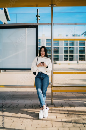 Photos Young woman with dental braces holding smartphone and looking away, career growth, ambition, successful digital nomad waiting at city bus stop, wearing white sweater and jeans, urban lifestyle