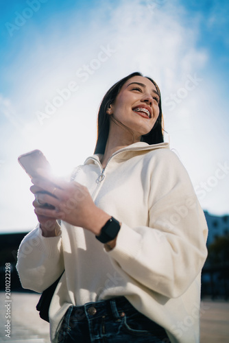 Smiling female with dental braces using mobile phone app while sitting at bus station, positive attitude, checking notifications, social media influence, urban background with glass walls.
