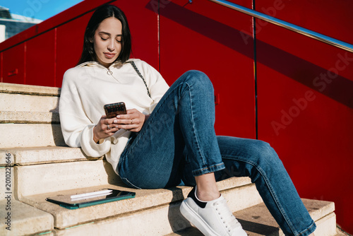 Young lady wearing earbuds and holding mobile phone while waiting for commute, listening to audio book or podcast, urban travel, serenity, mindfulness, contemporary digital lifestyle, city day.