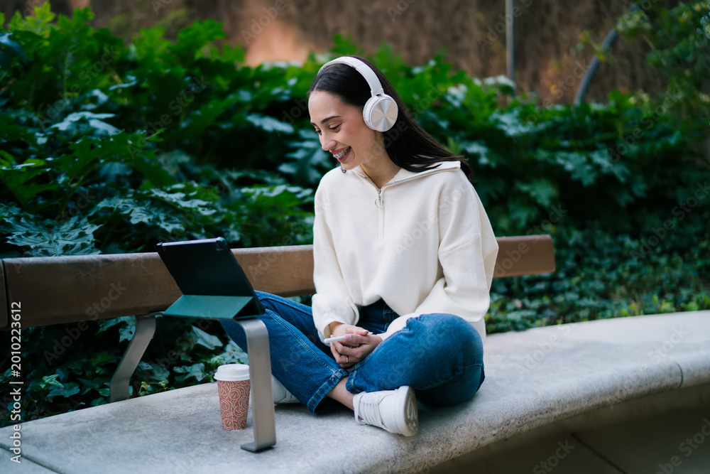 Fototapeta premium Cheerful girl with braces in wireless headphones sitting cross-legged on bench and using tablet. Happy woman enjoying audio content or video call in urban green park, digital nomad representation.