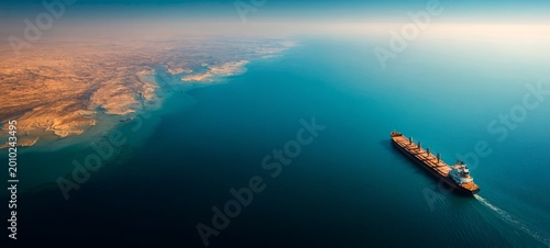 Massive Oil Tanker Crossing the Strait of Hormuz Under Bright Daylight, Calm Blue Sea and Desert Mountains, Symbolizing Global Energy Transport and Crude Oil Trade, Cinematic Wide Composition with Cop