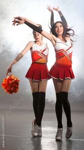Two Cheerful Young Asian Women Cheerleaders In Red And White Uniforms Jumping And Dancing With Orange Pom Poms In A Smoky Studio Environment