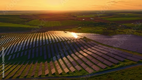 Rows of solar panels spread across a hillside near water. The sun sets in the background, casting warm light over the land. Fields and a rural area extend in the distance.