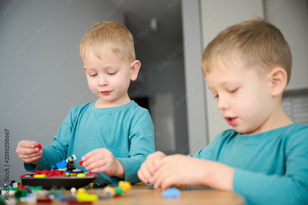 Fototapeta premium Two boys playing with building blocks, assembling colorful pieces and creating model. Sitting at table at home, creative play, learning and sibling interaction