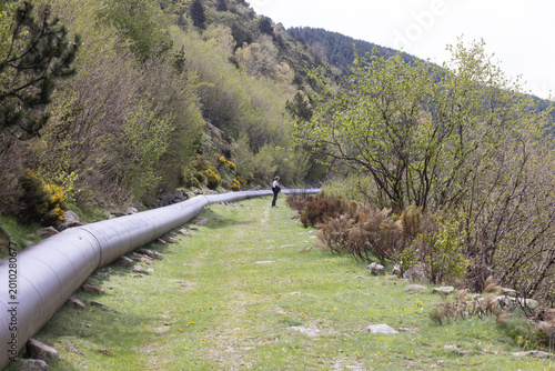 Une personne se trouve près d'une conduite forcée métallique posé sur une étroite bande de terre herbeuse, entourée de végétation et de collines boisées.