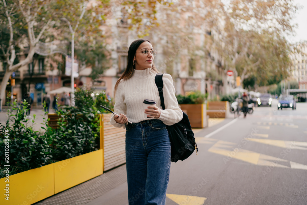 Fototapeta premium Attractive millennial girl walking city street holding smartphone and coffee cup, young woman in stylish white sweater and backpack looking away, urban youth lifestyle and digital nomad concept.