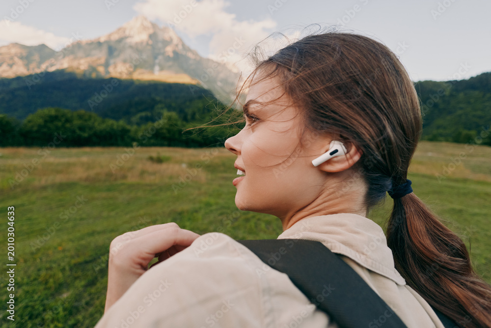 Naklejka premium Young woman with ponytail wearing wireless earbuds hiking in green meadow with mountain background enjoying nature and active lifestyle outdoors in fresh air.