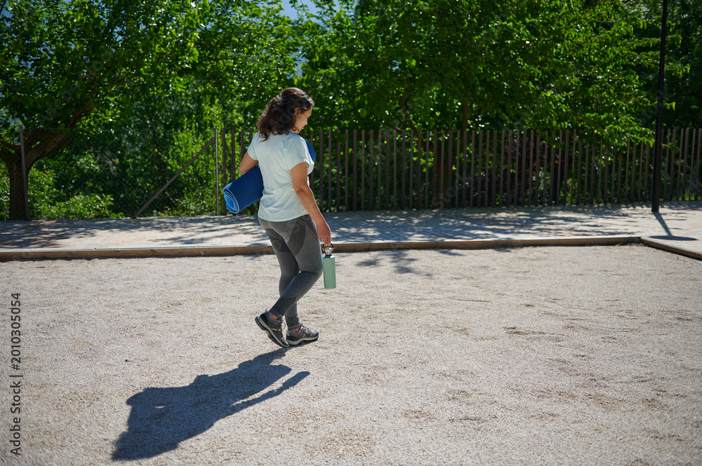 Fototapeta premium Woman Carrying Yoga Mat and Water Bottle Walking in Park After Workout