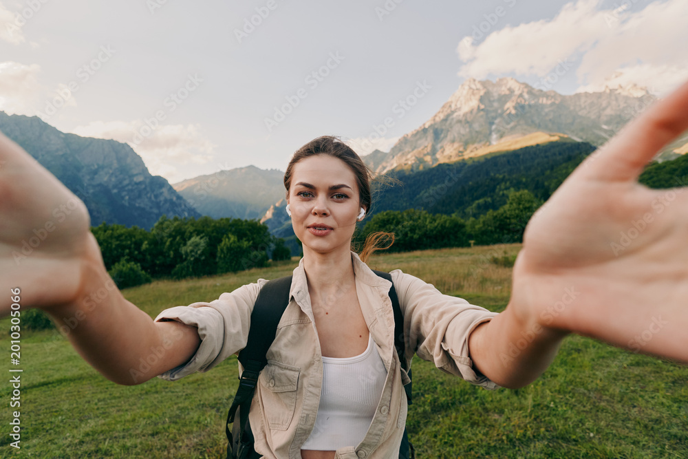 Naklejka premium A young woman in casual clothes takes a selfie outdoors in a green meadow with mountains in the background, enjoying nature and an active lifestyle.