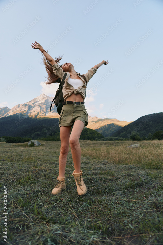 Naklejka premium Joyful woman jumping in a grassy field with mountains in the background, depicting outdoor lifestyle, freedom, and carefree adventure under a clear sky.