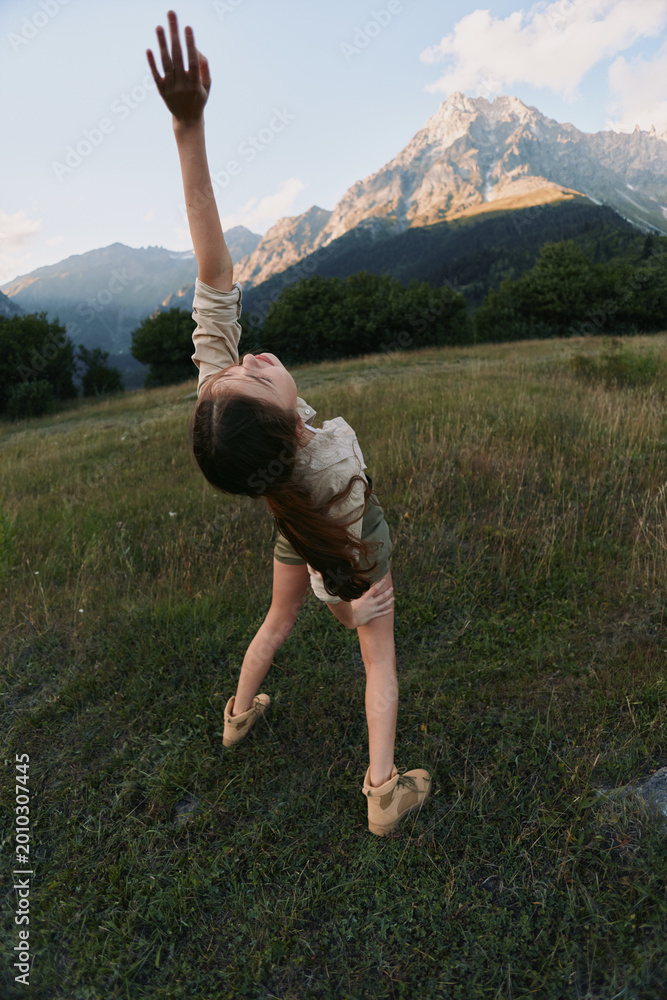Naklejka premium A young woman dances in a grassy meadow, arms raised toward the sky, mountains in the background, conveying freedom, vitality, and a carefree outdoor lifestyle.